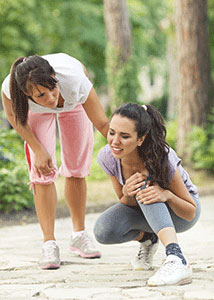 A girl knealing down and holding her hurt knee while her friend tries to comfort her.